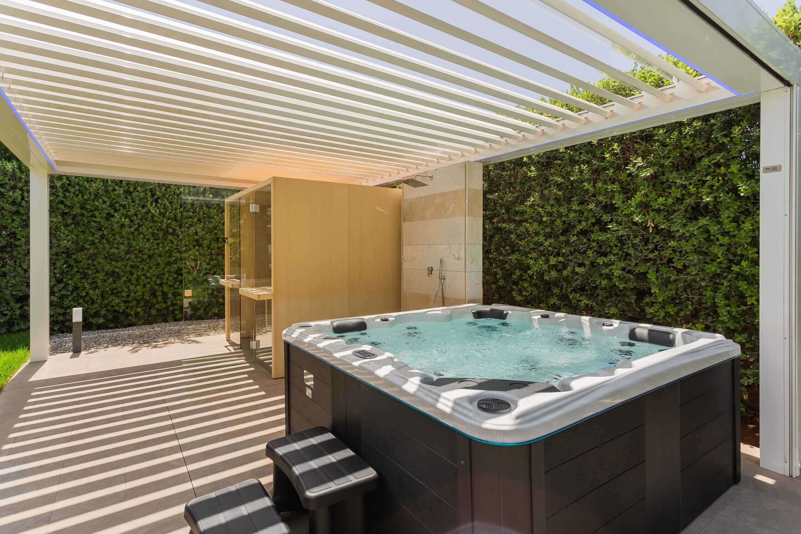 A modern outdoor hot tub area with a pergola overhead, a wooden cabinet, and a glass shower enclosure.