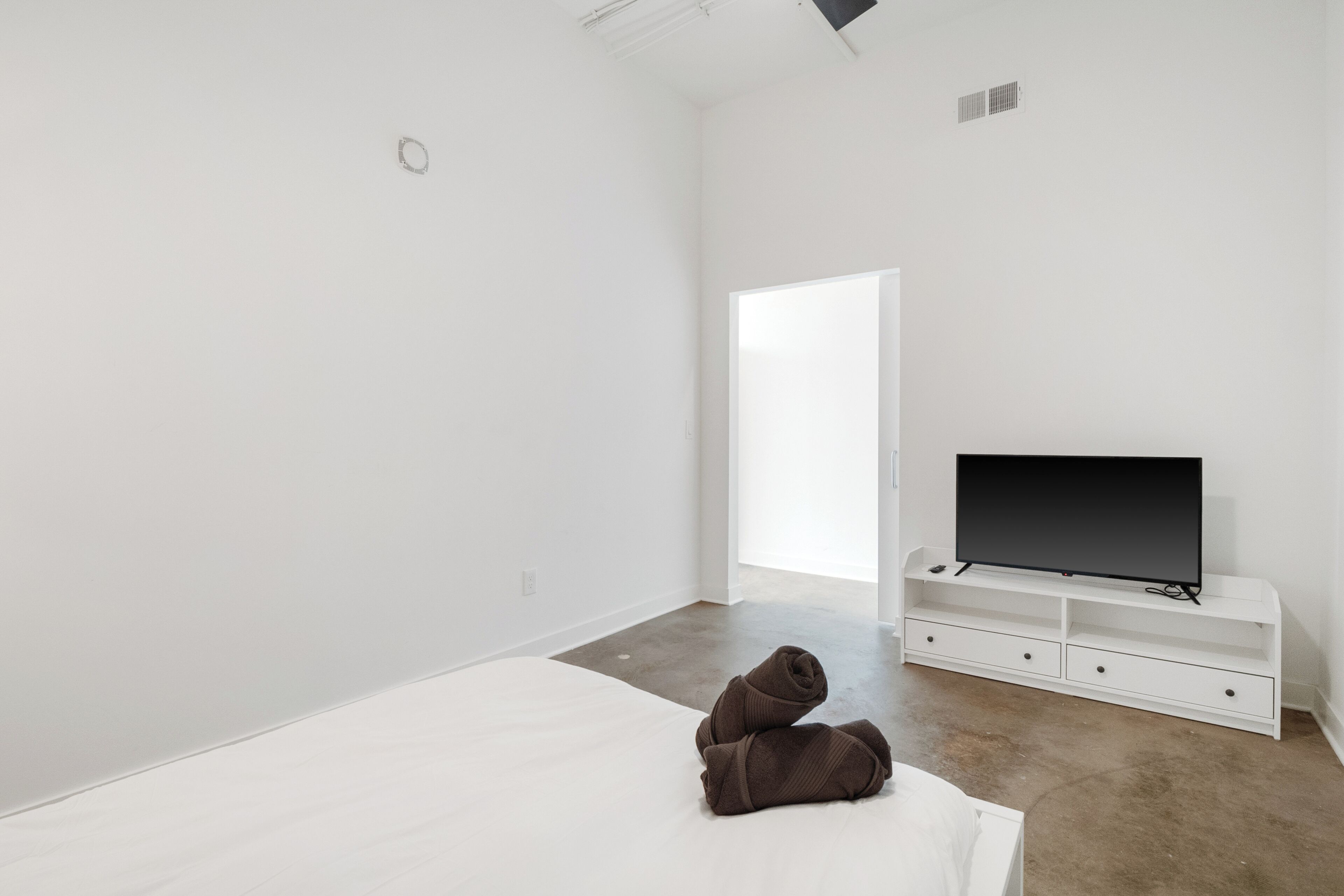 A minimalist bedroom with a bed, a TV on a white stand, and a door leading to another room.