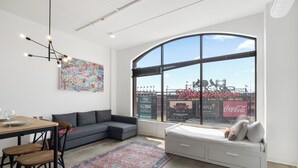 A modern living room with a large window, a sofa, a dining area with chairs, and a small bed. - Ballpark Luxury Loft Steps from Arch (St. Louis)