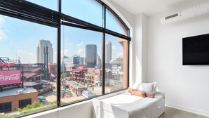 A modern bedroom with a large window offering a city view, a white bed, and a mounted TV. - Ballpark Luxury Loft Steps from Arch (St. Louis)