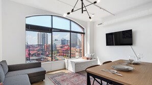 A modern living room with a large window, a flat-screen TV, a dining table set for two, and a cozy seating area. - Ballpark Luxury Loft Steps from Arch (St. Louis)