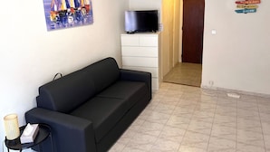 Un salon avec un canapé noir, un tableau représentant des voiliers, un téléviseur et un couloir menant à une porte.
