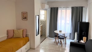 Une petite pièce bien éclairée, avec un lit, une télévision, une petite table avec des chaises et une fenêtre avec des rideaux.