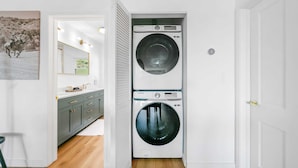 A laundry room with two stacked washing machines, a mirror, and a door leading to another room.