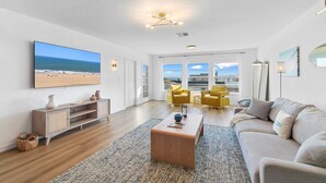 A spacious living room with a large TV, wooden flooring, a sofa, yellow chairs, and a view of the beach through large windows.