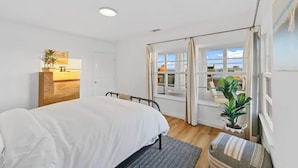 A bedroom with a bed, a dresser, a window with curtains, a plant, and a view of the outside.