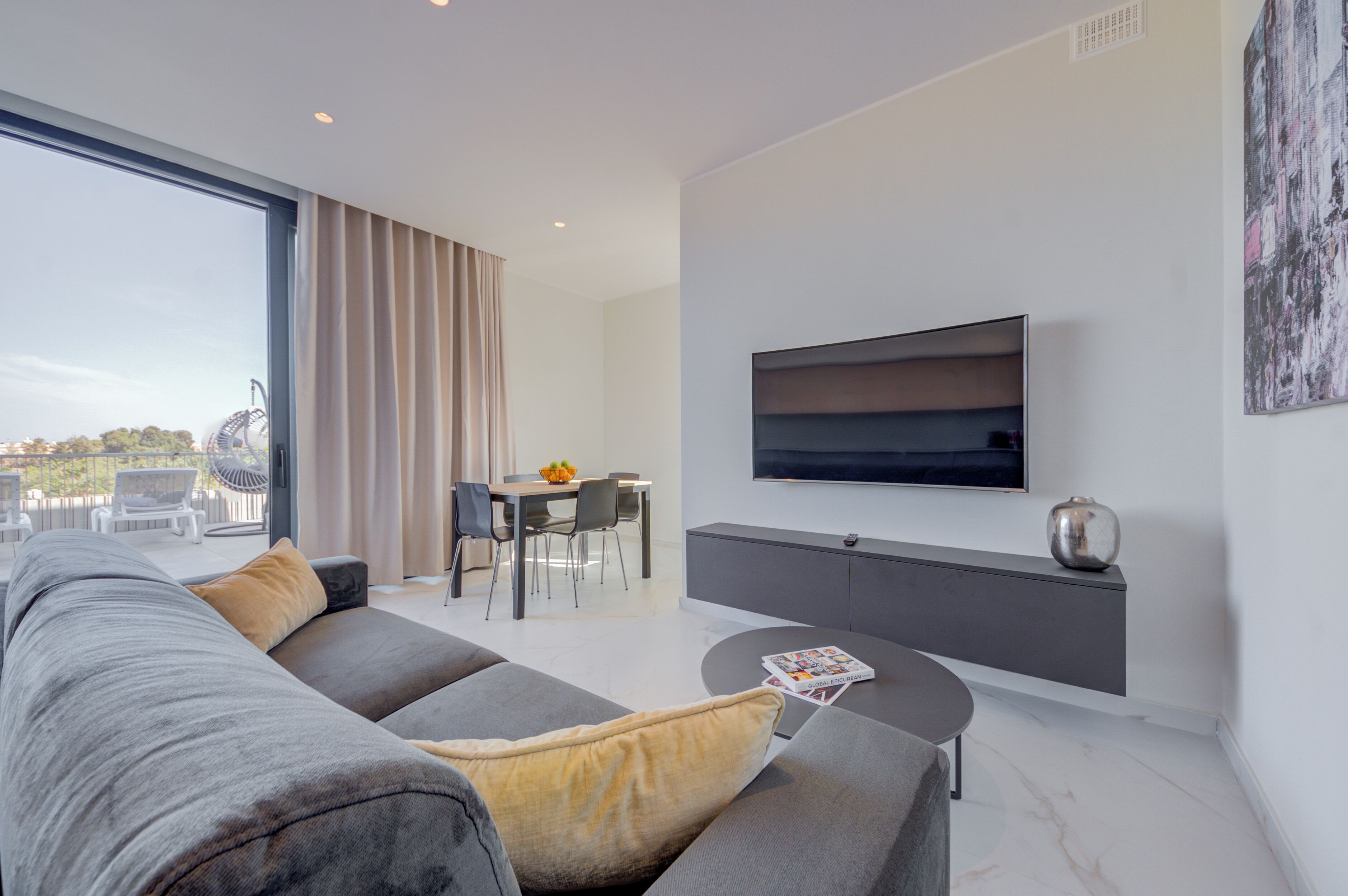 Un salon moderne avec un canapé gris, une table à manger avec des chaises, un téléviseur fixé au mur et un balcon offrant une vue sur des montagnes russes.