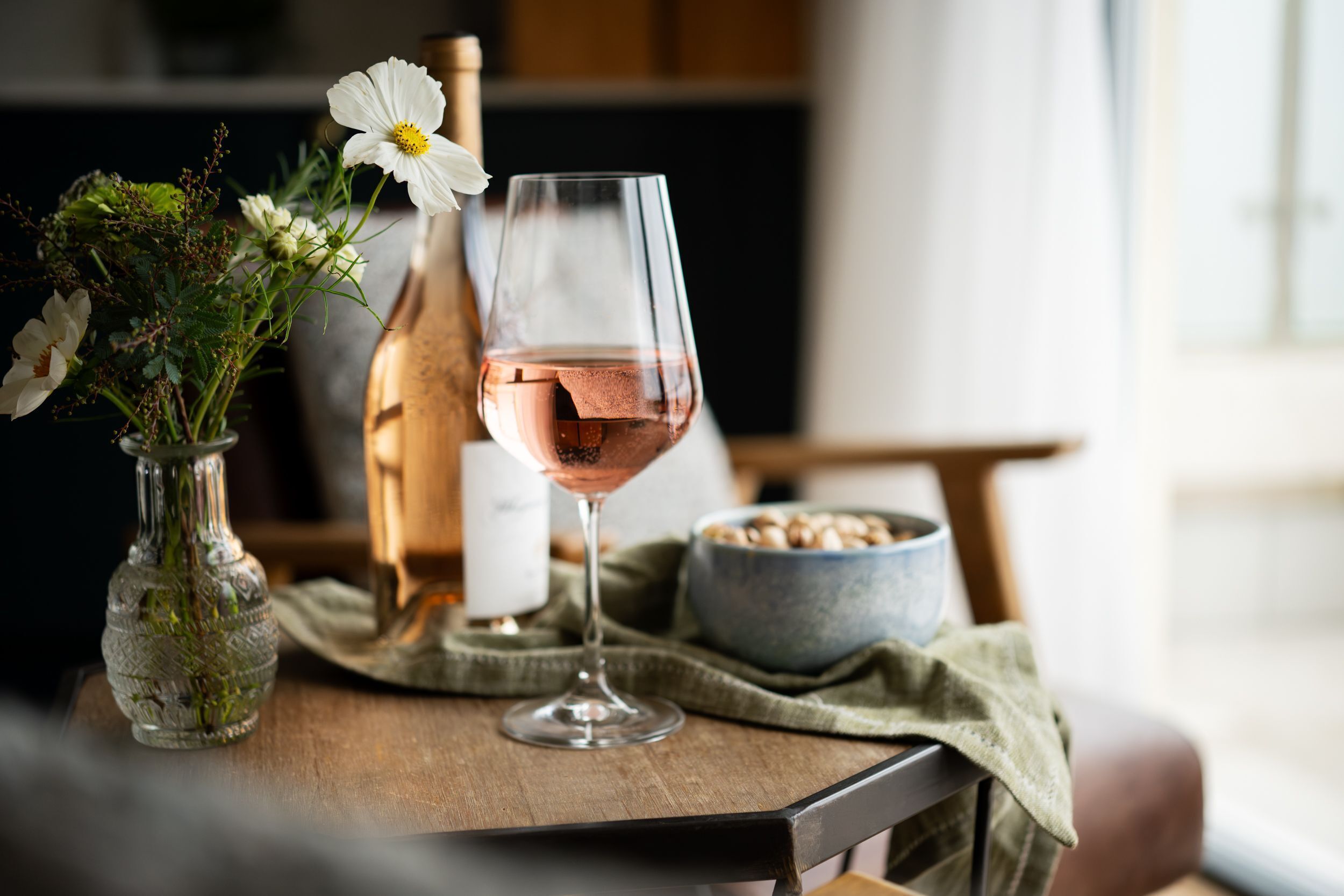 A table with a glass of rosé wine, a vase with flowers, and a bottle.