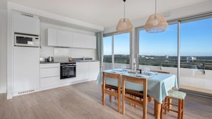 Une cuisine moderne avec des meubles blancs, un four à micro-ondes et une cuisinière. On y trouve un coin repas avec une table dressée pour deux personnes, ainsi que de grandes fenêtres offrant une vue sur la ville.