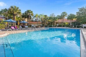 Uma piscina com espreguiçadeiras e guarda-sóis, cercada por palmeiras e um edifício com telhado vermelho.