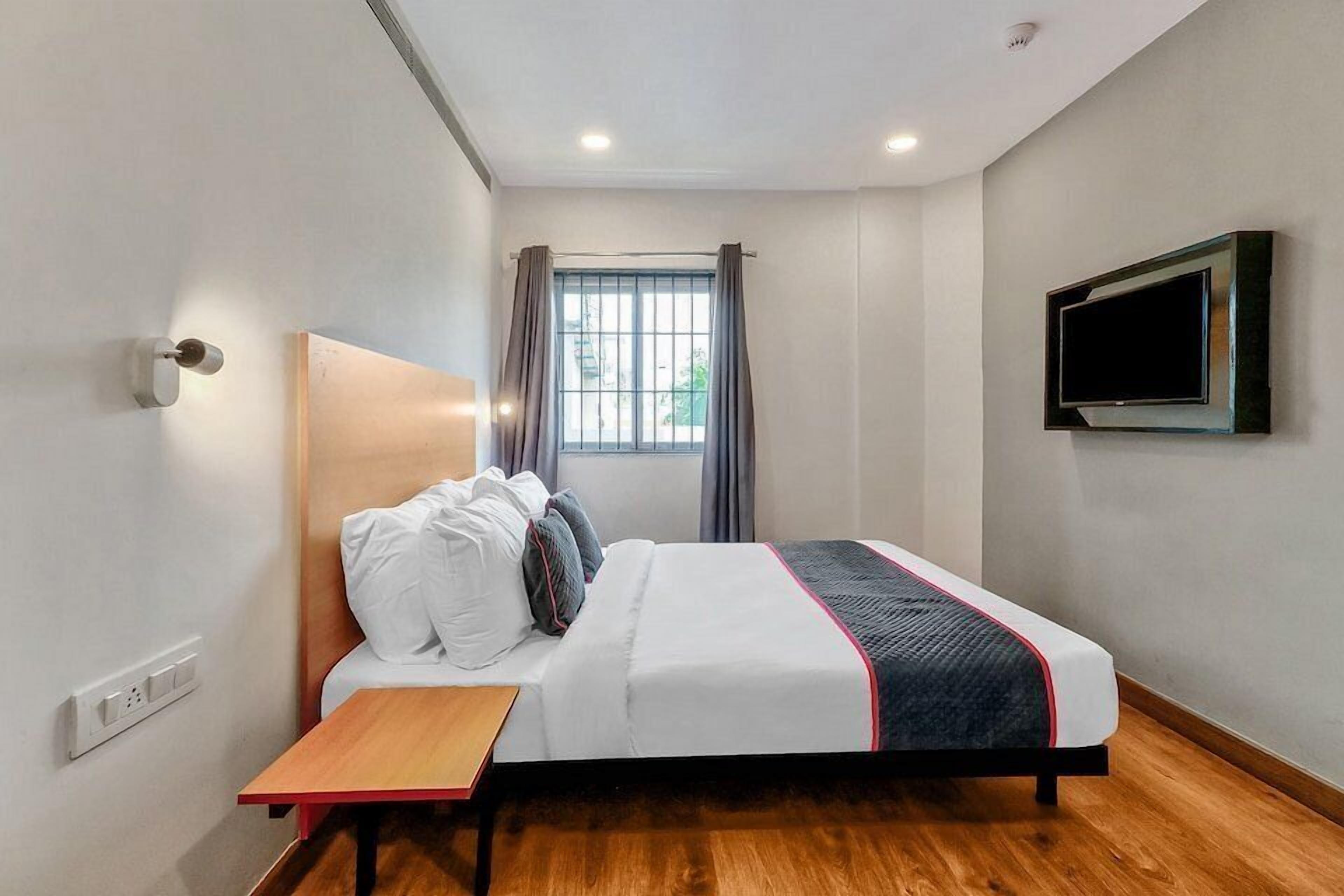 A hotel room with a bed, a TV mounted on the wall, a window with curtains, and a wooden floor.