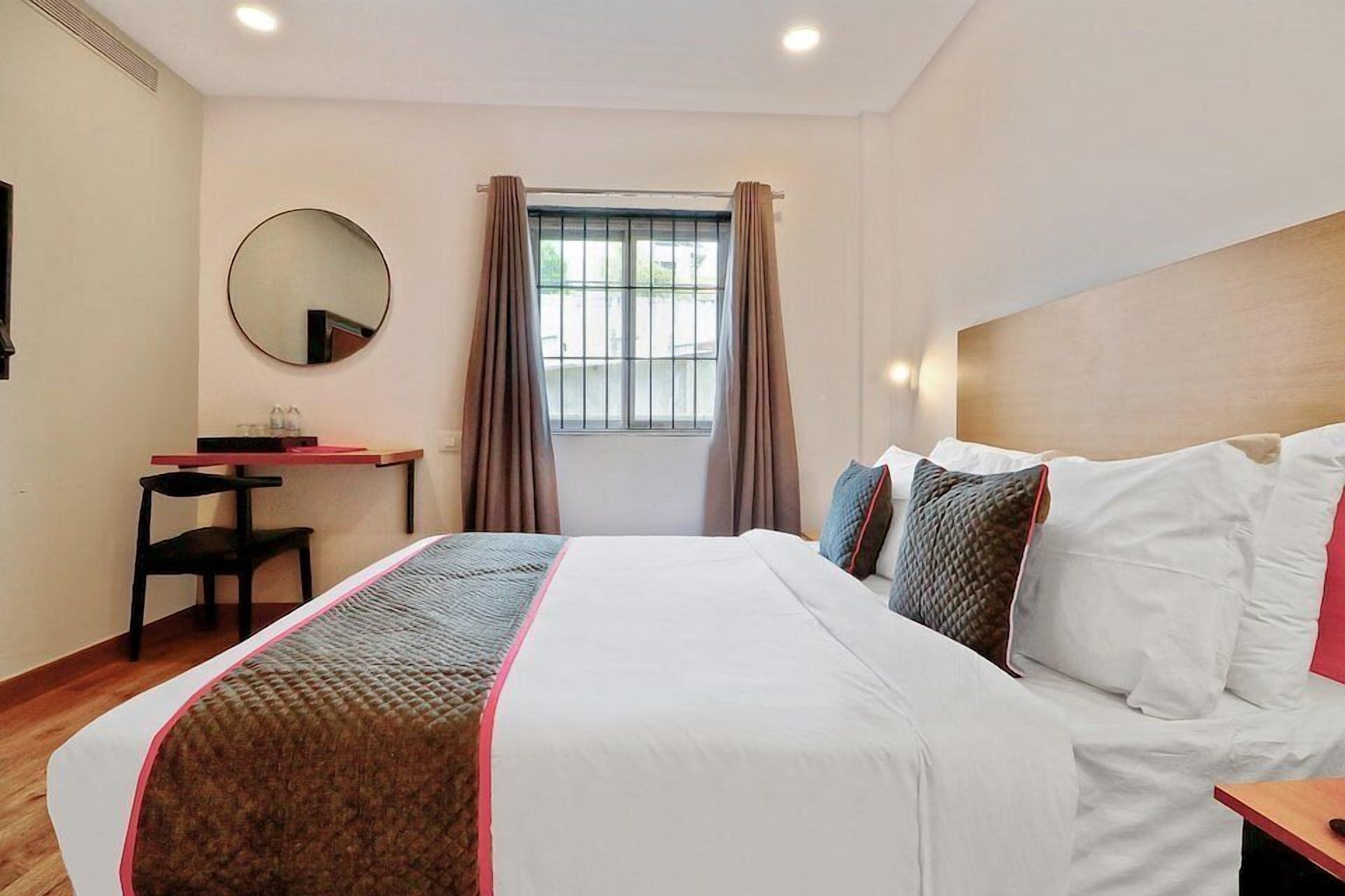 A hotel room with a bed, a desk, a chair, a round mirror, and a window with curtains.