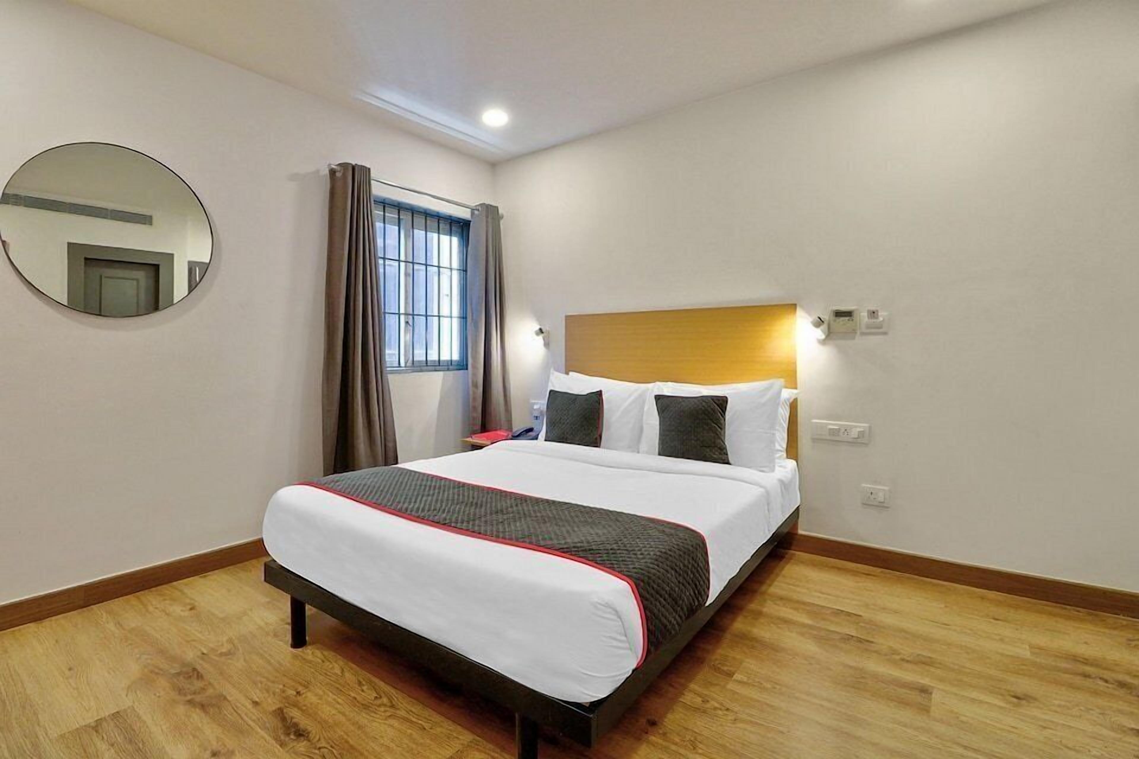 A hotel room with a bed, a round mirror, a window with curtains, and a wooden floor.