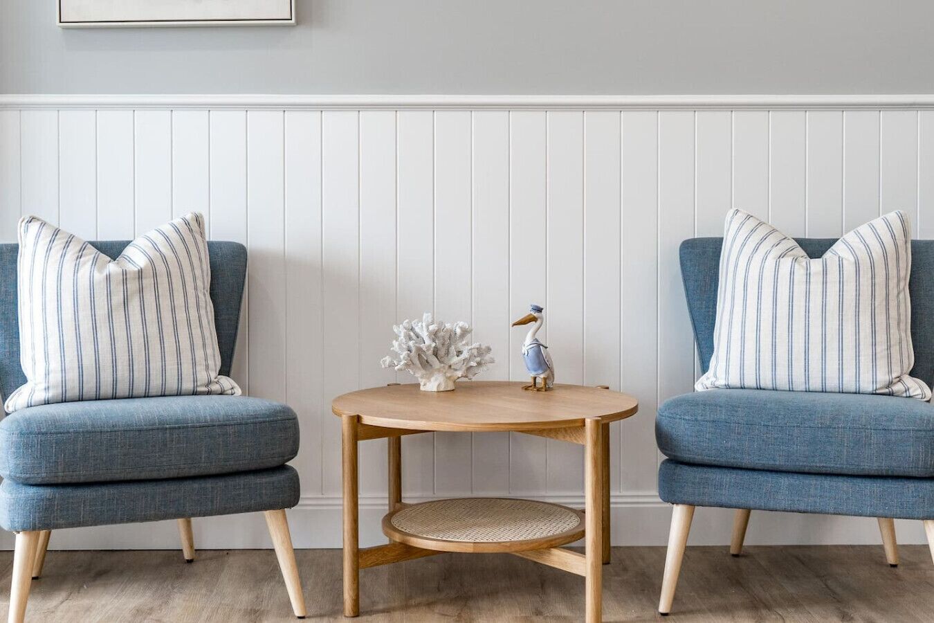 A room with two blue armchairs with striped pillows, a small wooden table with a woven chair, and decorative items including a coral sculpture and a seagull figurine.
