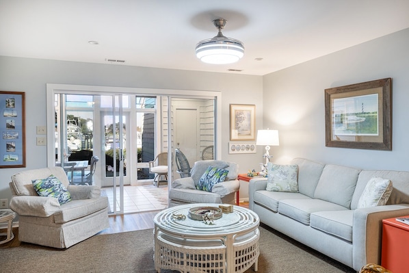 A living room with a white sofa, two armchairs, a coffee table, and a ceiling fan. There is a door leading to a patio area with outdoor furniture.