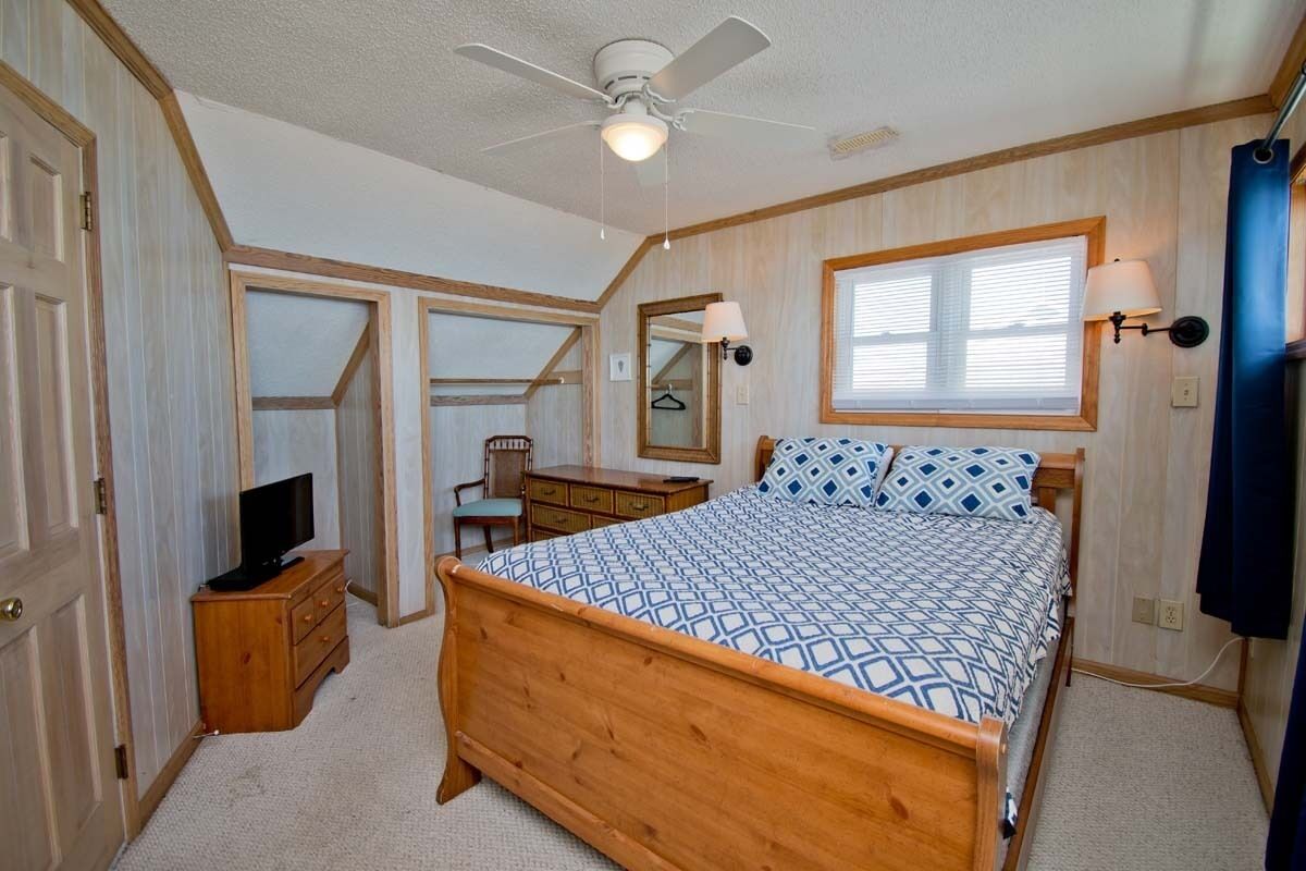 Een slaapkamer met een houten bed, een plafondventilator, een bureau met stoel, een televisie en een raam met jaloezieën.