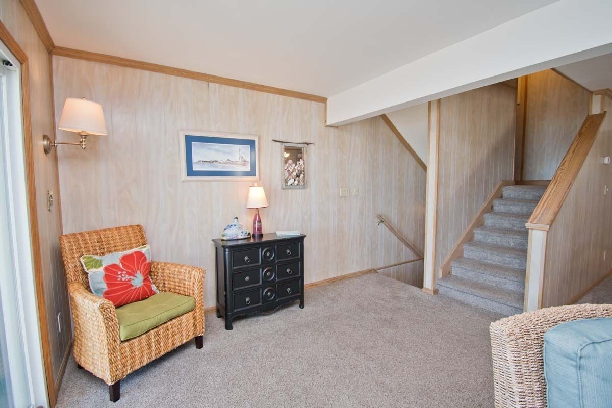 A room with a wicker chair, a black dresser, a framed picture, and a staircase.