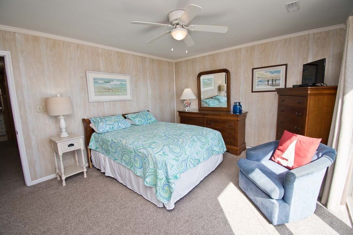 Ein Schlafzimmer mit Bett, Stuhl, Kommode, Spiegel und Deckenventilator.