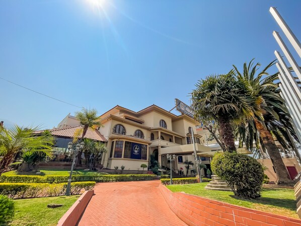 A two-story building with a tiled roof, a landscaped garden with hedges and palm trees, and a brick pathway leading to the entrance.