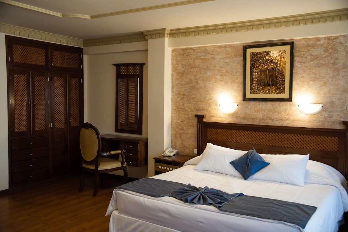 A hotel room with a large bed, a desk, and a chair. There is a telephone on the desk and a framed picture on the wall.