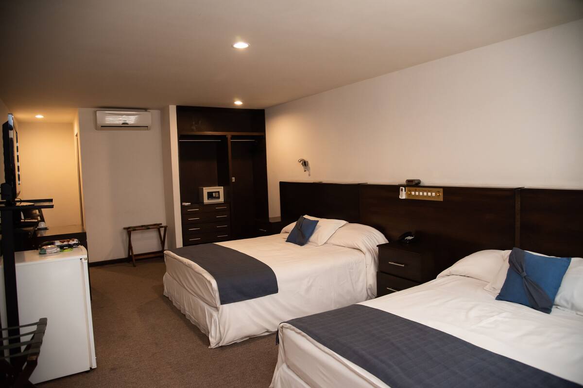 A hotel room with two beds, a desk, a refrigerator, a wardrobe, and an air conditioning unit.