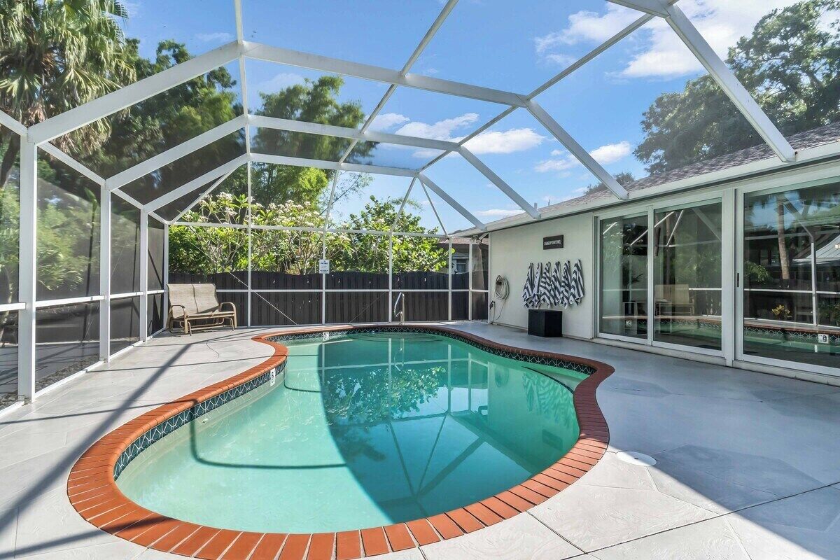 A covered swimming pool with a brick edge, surrounded by a glass enclosure, and a patio area with a chair.