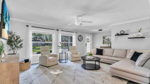A modern living room with a beige sofa, two armchairs, a coffee table, and a ceiling fan. Large windows allow natural light and provide views of the outdoors.