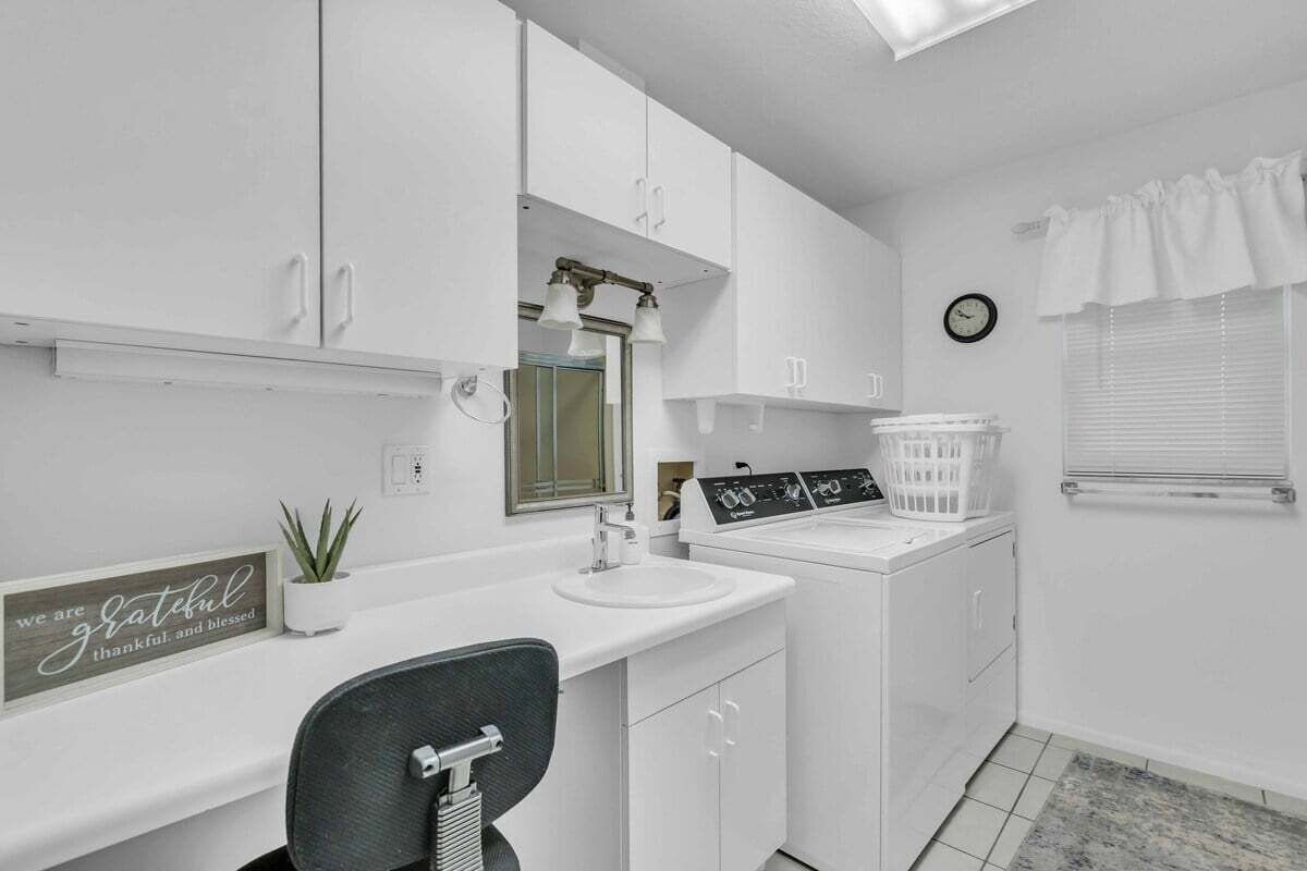 A modern bathroom with white cabinetry, a sink, a mirror, a clock, and a window with blinds.