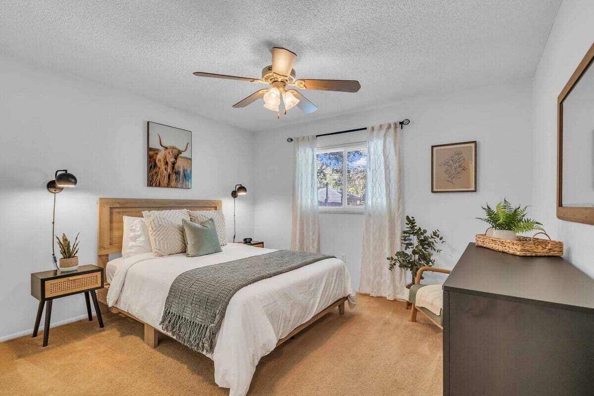A bedroom with a bed, bedside tables, a ceiling fan, a window with curtains, and wall art.