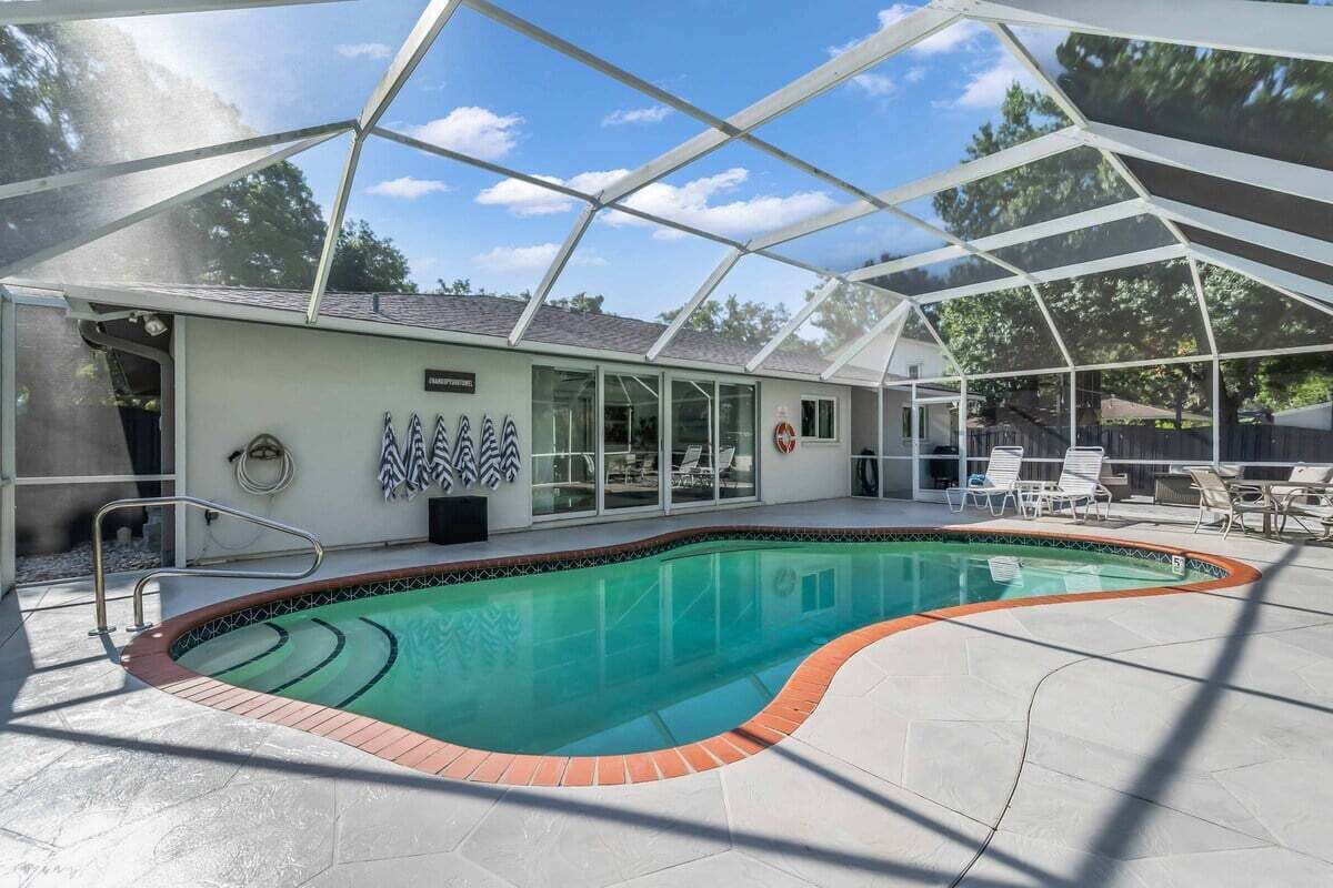A covered swimming pool with a glass enclosure, a tiled deck, and a lounge area with chairs.