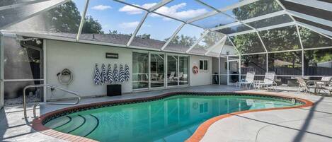 A covered swimming pool with a glass enclosure, a tiled deck, and a lounge area with chairs.