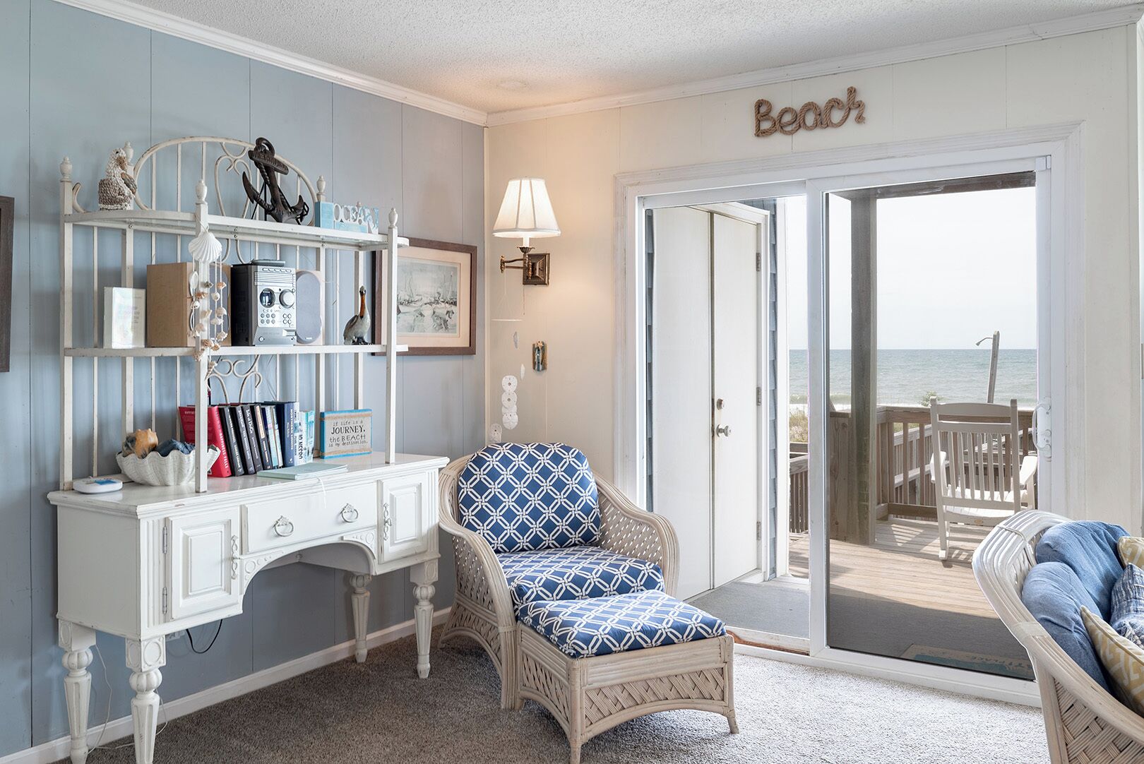A room with a white desk, a wicker chair with patterned cushions, a bookshelf, and a door leading to a balcony with a view of the sea.