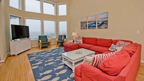 A spacious living room with a large red sofa, a television on a white cabinet, and a view of the sea through large windows. - Witt's Inn, Soundfront House in Emerald Isle, NC! (Emerald Isle)
