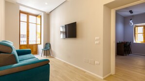 A modern living room with a teal sofa, a flat-screen TV mounted on the wall, and a dining area visible through an open door.