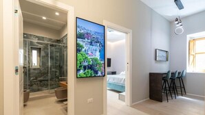 A modern interior with a shower area, a mounted TV, a dining area with chairs, and a bed in the background.