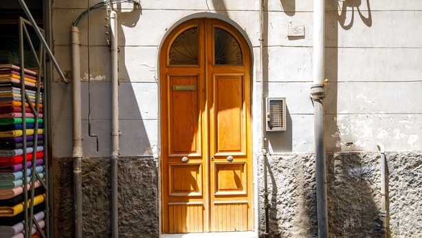 A wooden double door with a small window above it, set in a stone wall with pipes and a metal grate nearby.