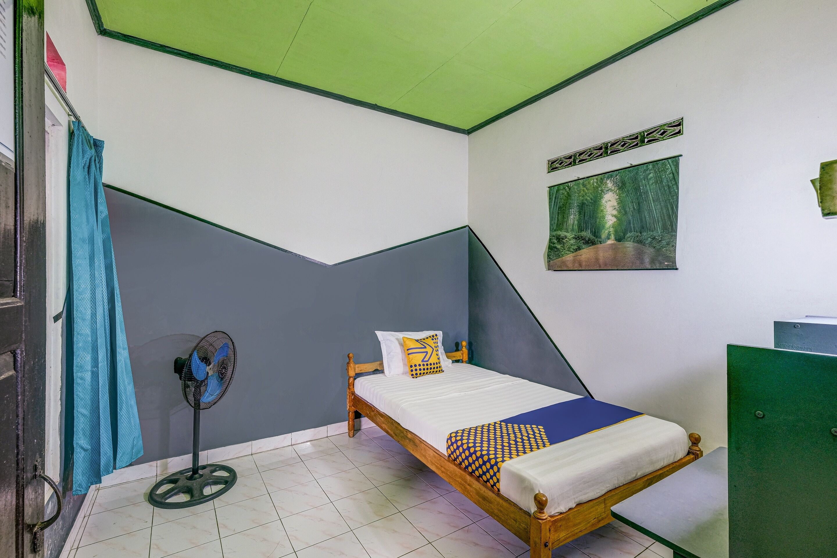 Una habitación con cama, ventilador de pie, una puerta verde y un cuadro en la pared.