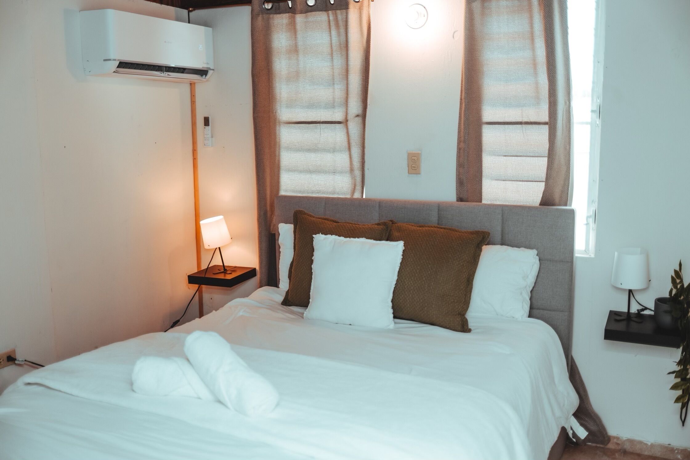 Una cama bien hecha con sábanas blancas y almohadas marrones, una mesita de noche con una lámpara, y una ventana con cortinas.