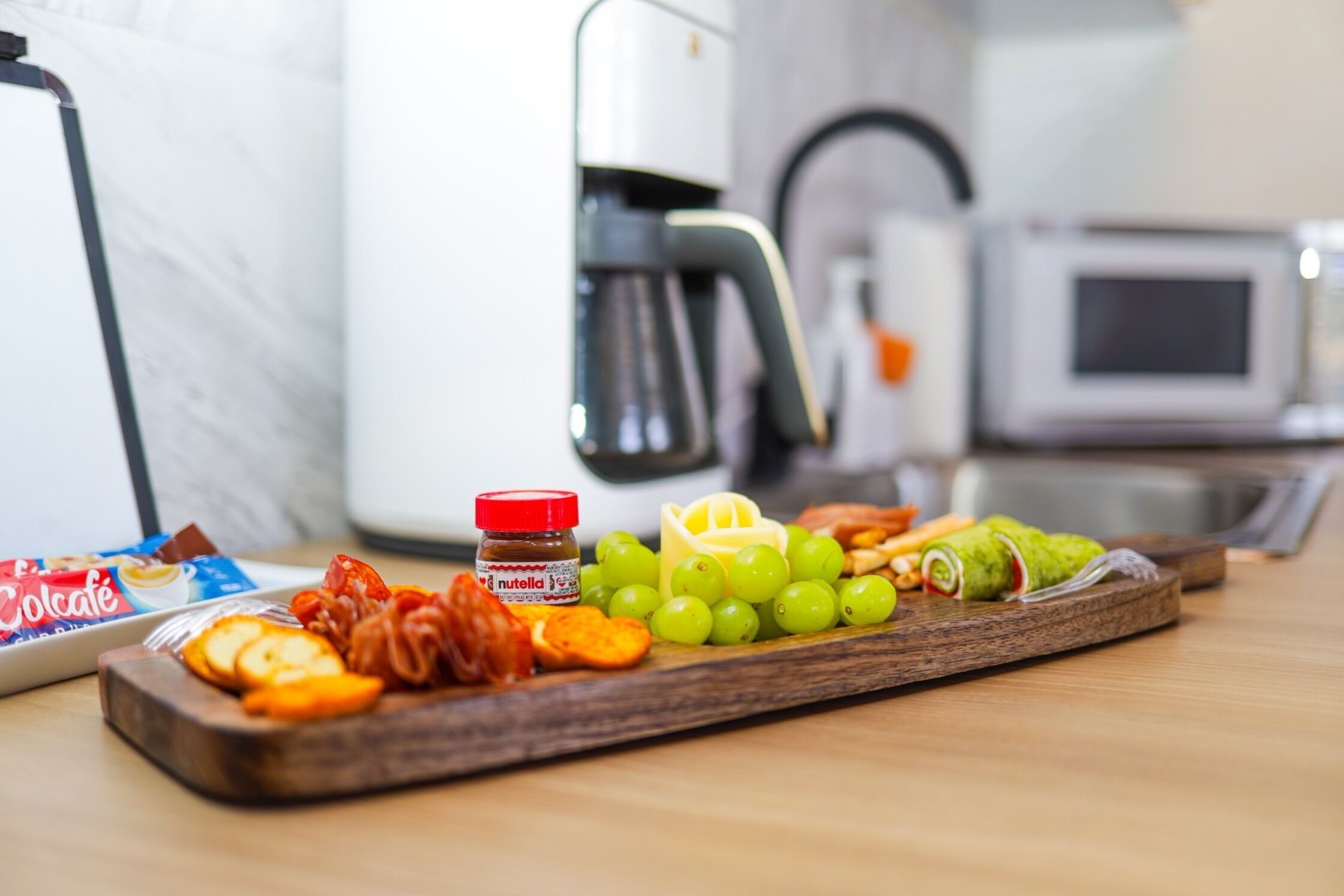 A wooden tray with snacks and a jar of Nutella, a microwave, and a coffee maker in a kitchen.