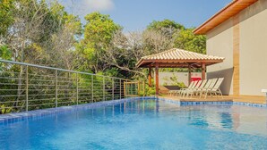 Una piscina con un quiosco de madera y sillas de descanso, rodeada por una cerca metálica y árboles.
