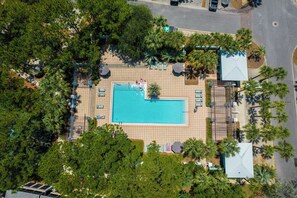 An aerial view of a resort with a central swimming pool, surrounded by sun loungers and umbrellas, with palm trees and a road nearby.