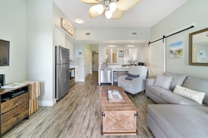 A spacious living area with a grey sofa, a wooden coffee table, and a kitchen with white cabinets and a refrigerator.