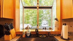 Una cocina con armarios de madera, una ventana con vista a árboles y un barco, un lavamanos y diversos utensilios de cocina.