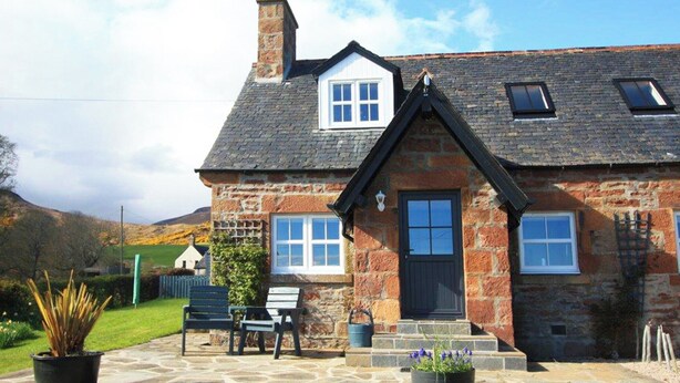 A stone house with a black door, white windows, and a stone staircase leading to the entrance.