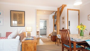 A cozy living room with a sofa, a wooden coffee table, a dining table with chairs, a staircase, and a kitchen in the background.