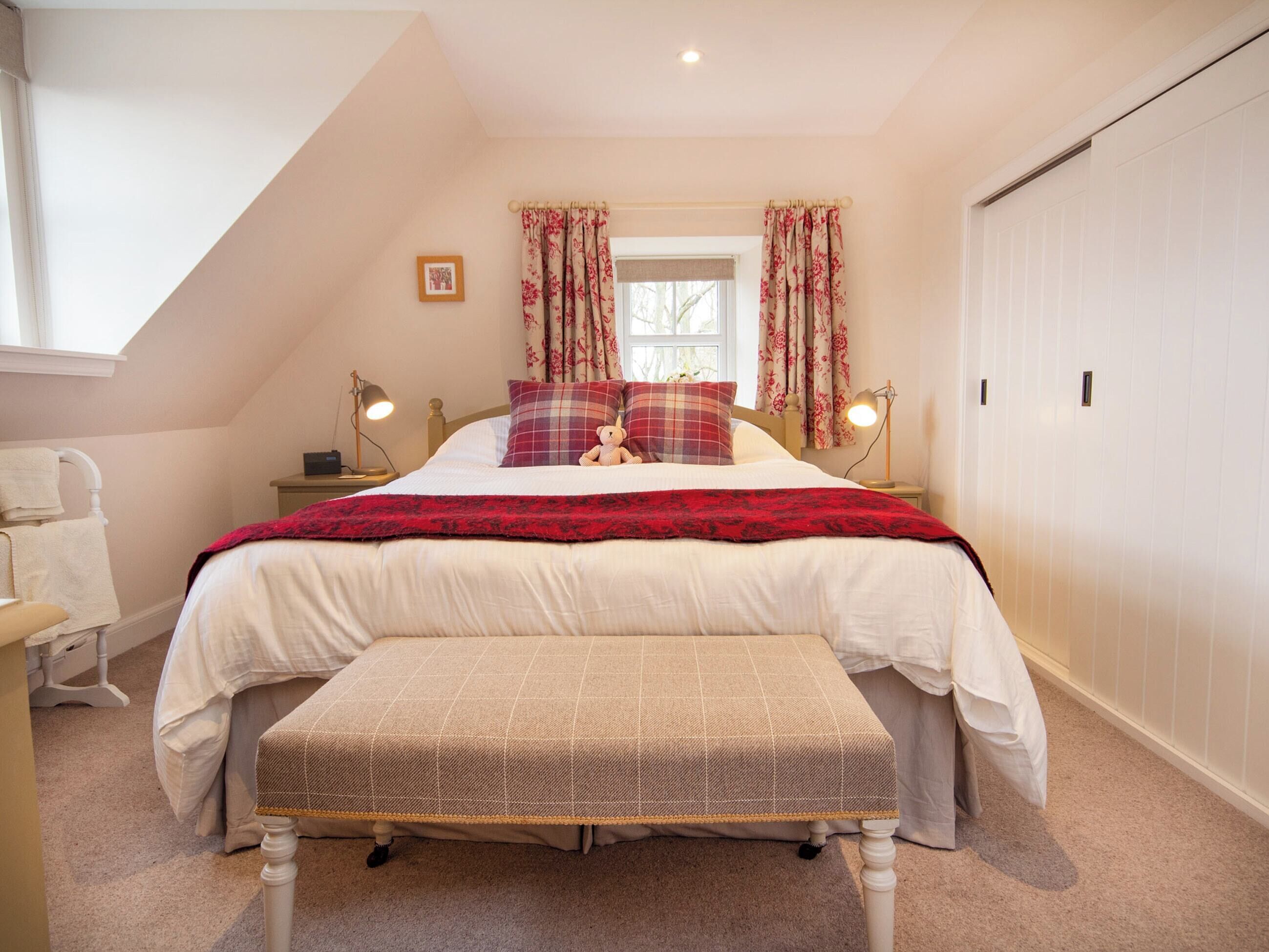 A bedroom with a bed, a bench, a nightstand, a lamp, a window with curtains, and a picture on the wall.