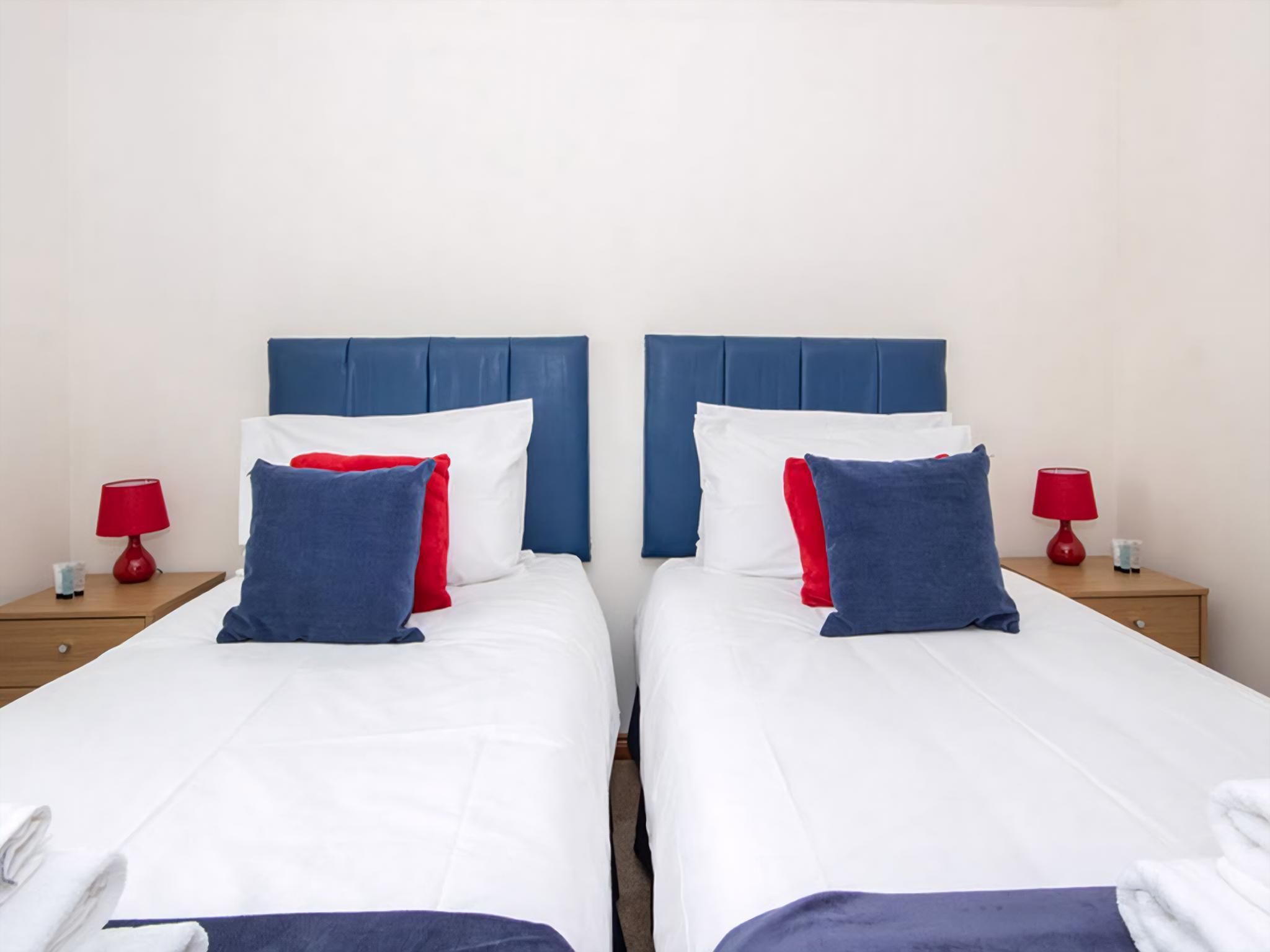 Habitación doble con dos camas, cada una con cabecera azul, ropa de cama blanca y almohadas rojas y azules. Hay dos mesitas de noche con lámparas rojas y un objeto pequeño en cada una.