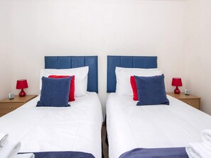 A double room with two beds, each with a blue headboard, white bedding, and red and blue pillows. There are two bedside tables with red lamps and a small item on each. - 2 bedroom accommodation in Inverness (Inverness)