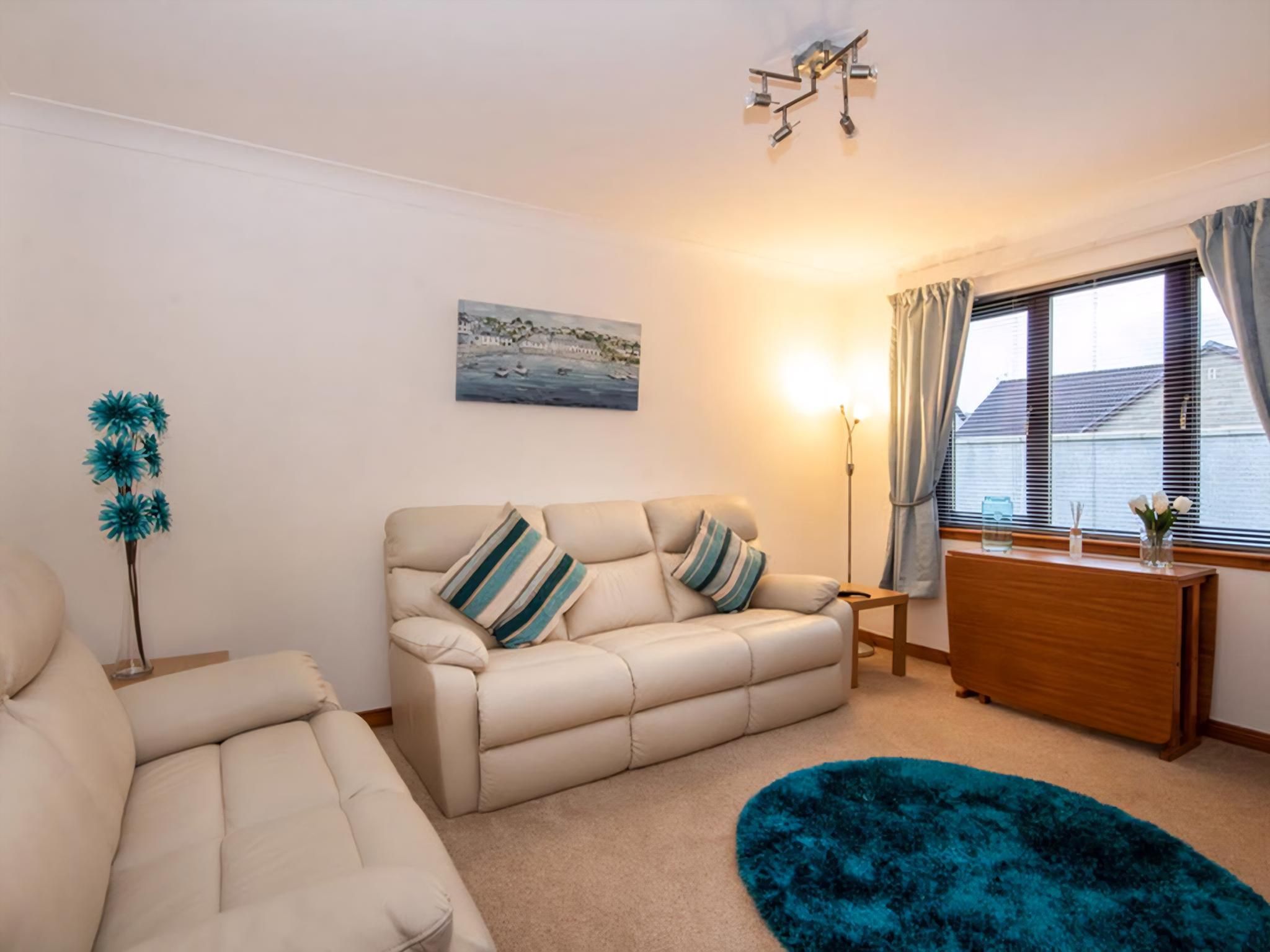 A living room with a beige sofa, a blue rug, a painting of a coastal scene, and a window with curtains.