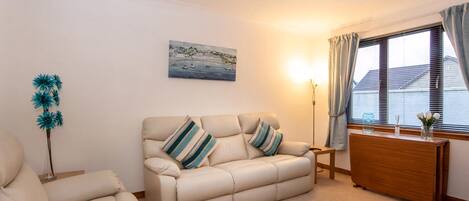 A living room with a beige sofa, a blue rug, a painting of a coastal scene, and a window with curtains.